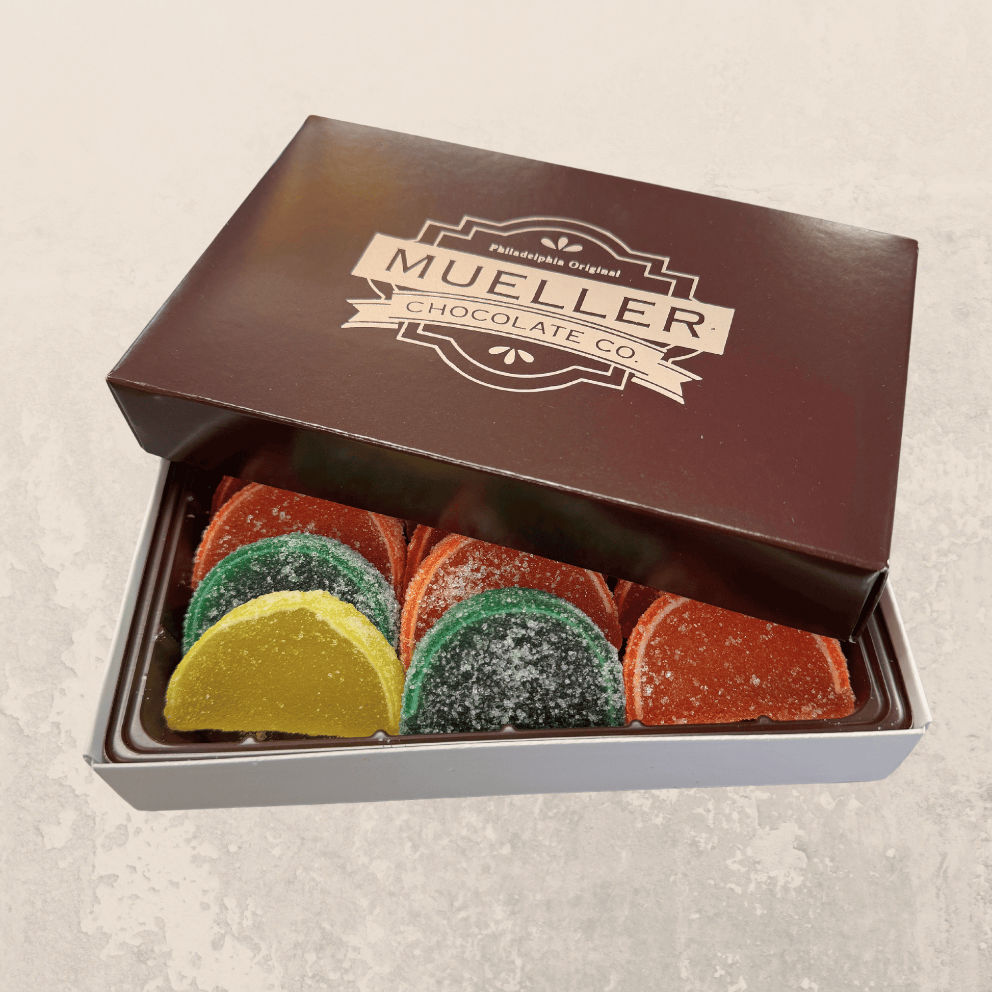 Mueller Chocolate Co. box of jellied fruit slices candy in orange, lemon, lime, and cherry flavors, coated in sugar and displayed on a metal surface.